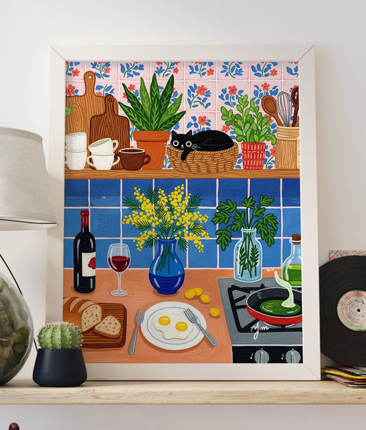 Whimsical kitchen illustration featuring a black cat curled up in a round wicker basket on a wooden shelf lined with stacked mugs, cutting boards, and potted houseplants. The wall is covered in hand‑painted pink tiles with blue and red floral motifs. Below, a blue tiled backsplash frames a cheerful countertop scene: a cobalt vase of yellow mimosa blossoms, fresh parsley in a glass jar, a bottle and glass of red wine, sliced artisan bread on a wooden board, a plate with two sunny‑side‑up eggs, scattered gold