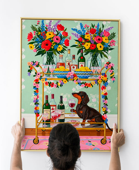 A whimsical illustration of a dachshund dog sitting beside a gold bar cart holding a glass of red wine. The cart is decorated with vibrant flowers and topped with wine bottles, lemon bowls, and two large floral bouquets. The background features green wallpaper with a light floral pattern and a pink rug underfoot. This colorful and joyful artwork combines humor, elegance, and charm, perfect for dog and wine lovers.