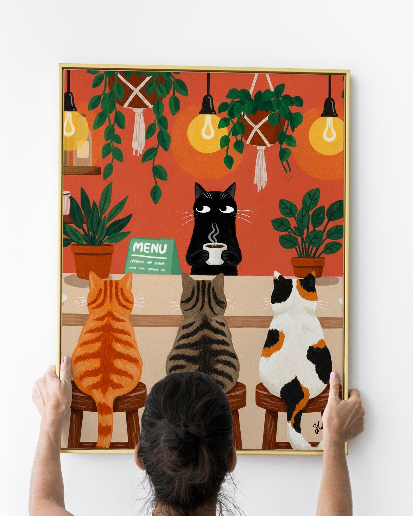 A whimsical illustration of a cozy cat café scene where three cats—a ginger tabby, a gray striped tabby, and a calico—sit on wooden stools at a coffee bar. A black cat barista stands behind the counter holding a steaming cup of coffee. The café is warmly lit with glowing hanging bulbs and decorated with lush green potted and hanging plants. The background features an earthy orange-red wall, creating a comforting and inviting atmosphere.